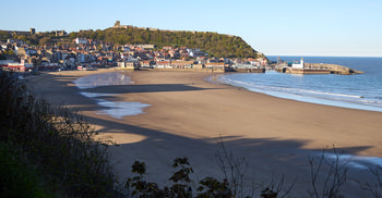 This landscape photograph captures Scarborough beach in the evening during spring, as evidenced by the lush greenery and the long shadows cast by the setting sun. The expansive sandy shoreline leads to the waters of the North Sea, with gentle waves approaching the coast. Above the town, Scarborough Castle stands prominently atop a hill, offering a historic landmark that overlooks the picturesque coastal scene. The town of Scarborough, located in Yorkshire, features a collection of traditional buildings clustered between the beach and the castle, creating a harmonious blend of coastal and urban elements. The peaceful ambiance and soft light of early evening give the beach a calm, inviting atmosphere, characteristic of Yorkshire’s scenic beauty.